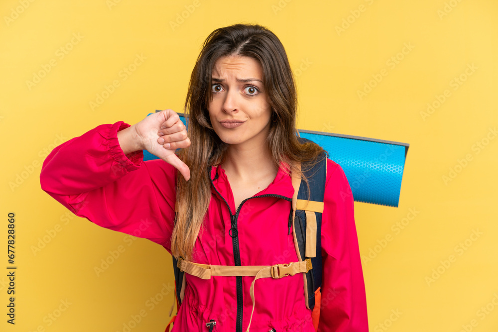 Young mountaineer man with a big backpack isolated on yellow background showing thumb down with negative expression Young mountaineer man with a big backpack isolated on yellow background showing thumb down with negative expression