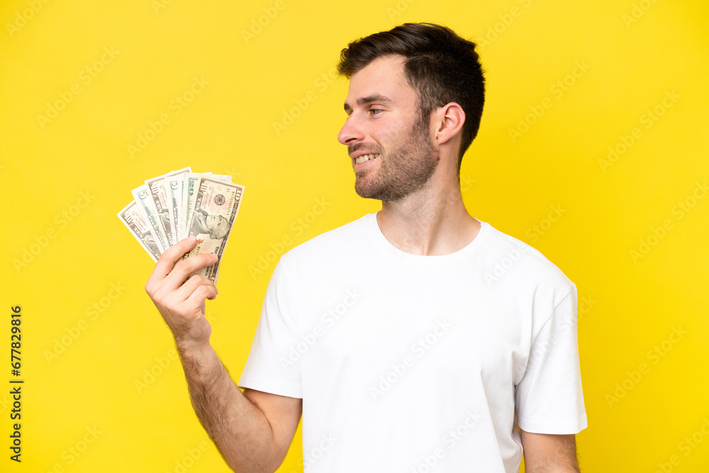 Young caucasian man taking a lot of money isolated on yellow background looking side Young caucasian man taking a lot of money isolated on yellow background looking side
