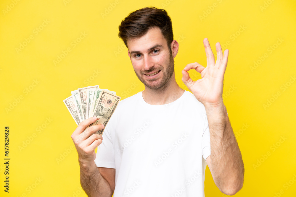 Young caucasian man taking a lot of money isolated on yellow background showing ok sign with fingers Young caucasian man taking a lot of money isolated on yellow background showing ok sign with fingers