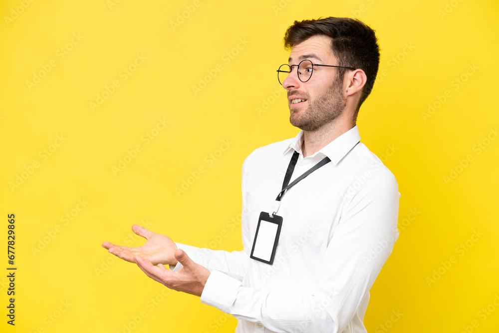 Young caucasian with ID card isolated on yellow background with surprise expression while looking side Young caucasian with ID card isolated on yellow background with surprise expression while looking side