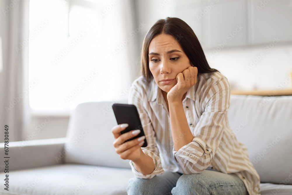 Upset Young Woman Sitting On Couch And Looking At Smartphone Screen Upset Young Woman Sitting On Couch And Looking At Smartphone Screen