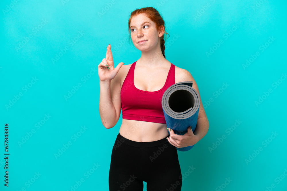 Young sport reddish woman going to yoga classes while holding a mat isolated on blue background with fingers crossing and wishing the best Young sport reddish woman going to yoga classes while holding a mat isolated on blue background with fingers crossing and wishing the best