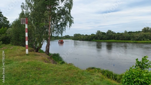 A wooden raft with a canopy floats down the river Shrubs and trees grow on the grassy banks of the river There are ripples on the water. On the shore is a wooden pole with a sign of the ship's passage photo