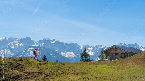 Auf dem Staffel Berg in den Bayerischen Voralpen läuft eine Wanderin zu einer Hütte mit schneebedeckten Bergen im Hintergrund.