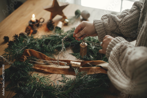 Hands in cozy sweater making Christmas rustic wreath with fir branches, ribbon, pine cones, bells on wooden table, close up. Winter holiday preparations, atmospheric time photo