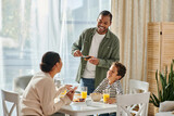 cheerful african american man spreading jam on toast and looking at his wife and son at breakfast