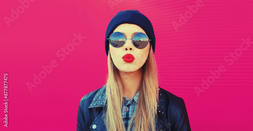 Portrait of stylish beautiful blonde woman blowing her red lips sending sweet air kiss wearing round sunglasses, black rock style, leather jacket and hat on pink background