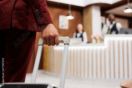 White collar worker with luggage arriving at hotel reception lobby, preparing to see room reservation. Young adult travelling on business meetings, carrying suitcase internationally. Close up. photo