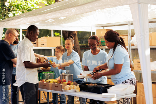 Individuals of diverse backgrounds gather outdoors to give donated food and non-perishables, providing support to the hungry and homeless. Multiracial volunteers share fresh free meals to poor people. photo