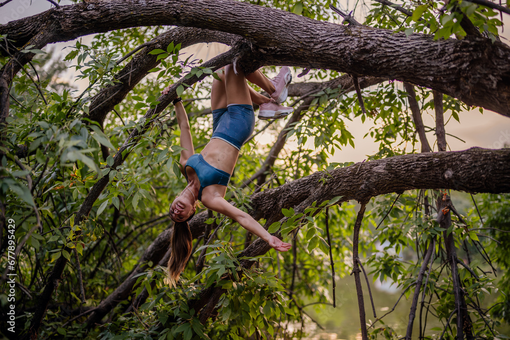 Fit girl with athletic body shape exercises outdoors. She climbs trees, showcasing strength and flexibility. Enjoying a green park environment, she prepares and warms up for training.