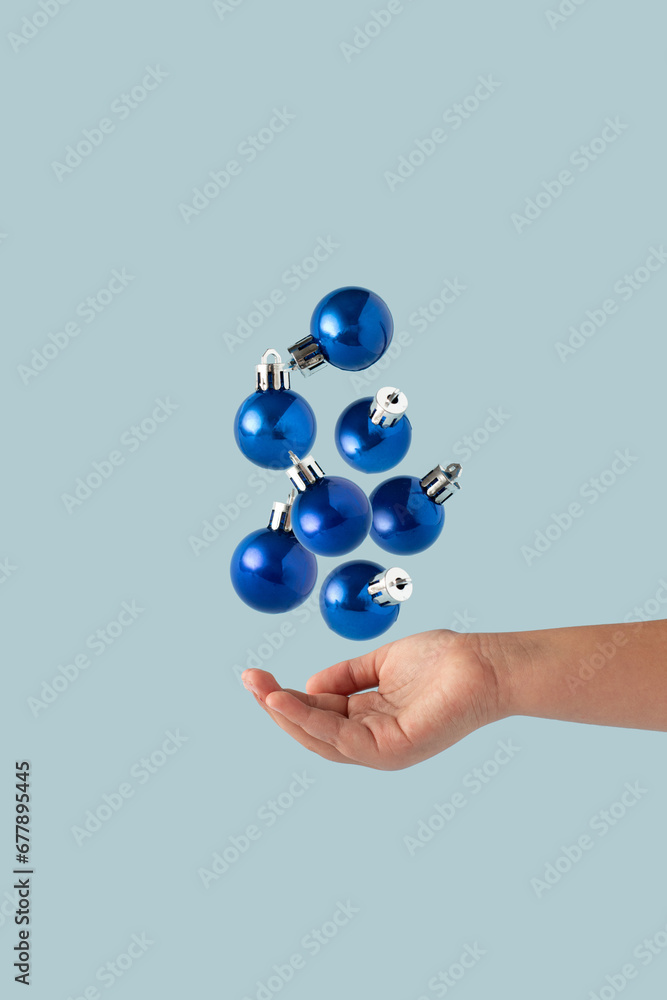 Fototapeta premium Shiny Blue Christmas decoration balls falling down into hand isolated on pastel blue background. Blue Christmas aesthetics.