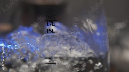 water boils bubbles in a transparent glass electric kettle close-up view. blue backlight in the dark, blurred background. divisions max min and water volume