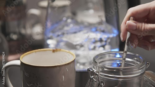 a young woman's hand pours a spoon of coffee from a transparent jar into a large cup on the background of a boiling kettle