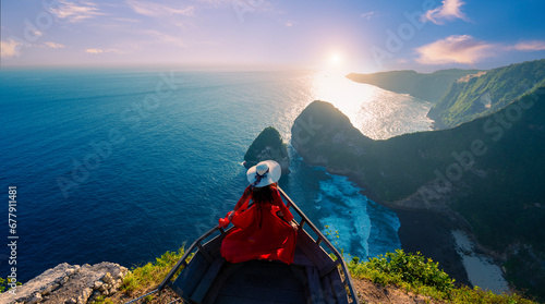Woman in red dress at Kelingking Beach viewpoint with the light of the sun shining at sunset On the island of Nusa Penida, Indonesia photo