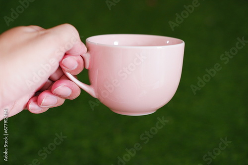 Female hand holding a pink cup of coffee on green grass background.