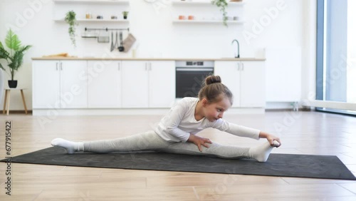 Fitness, little preschool female training yoga in half tortoise pose in kitchen floor at home, copy space. Young slim girl in white sportswear makes stretching training doing splits exercise.