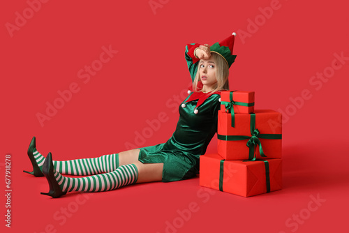 Tired woman in elf costume with Christmas gift boxes on red background photo