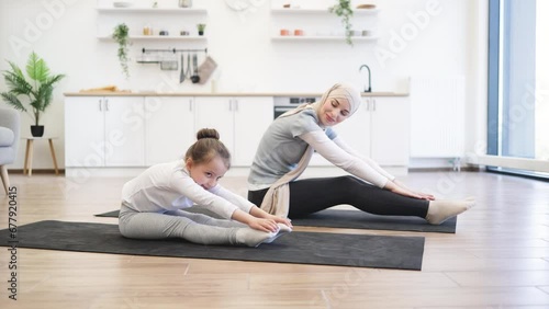 Young muslim mother in hijab showing her little daughter legs stretching exercises. Happy girls training at home. Motherhood and sporty lifestyle concept. photo