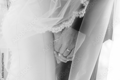 A black and white portrait of a bride and groom holding hands underneath a lace veil on their wedding day