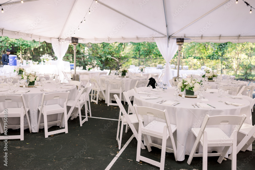 White tables and white chairs set up outside under a tent at an outdoor wedding reception White tables and white chairs set up outside under a tent at an outdoor wedding reception