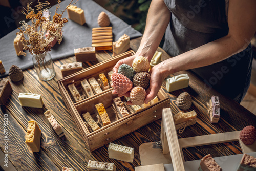 A woman soap maker holds soap in her hands in the form of colorful coniferous cones. A lot of different sliced pieces in a wooden box. Eco-friendly natural handmade cosmetics