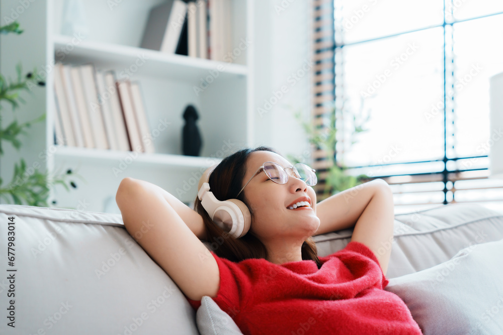 Relaxed young Asian woman enjoying rest on comfortable sofa, calm attractive girl relaxing on couch, breathing fresh air with eyes closed, wearing eyeglasses and headset, meditating at home Relaxed young Asian woman enjoying rest on comfortable sofa, calm attractive girl relaxing on couch, breathing fresh air with eyes closed, wearing eyeglasses and headset, meditating at home