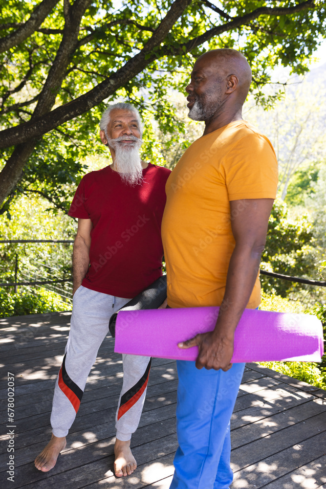 Obraz premium Two happy diverse male senior friends with mats talking before yoga class in sunny garden
