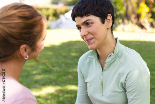 Happy biracial lesbian couple practicing yoga sitting and smiling at each other in sunny garden