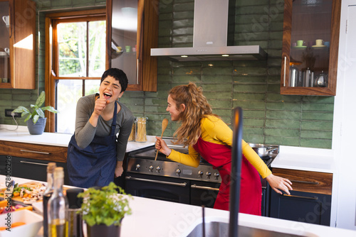 Happy biracial lesbian couple preparing meal having fun singing in kitchen photo
