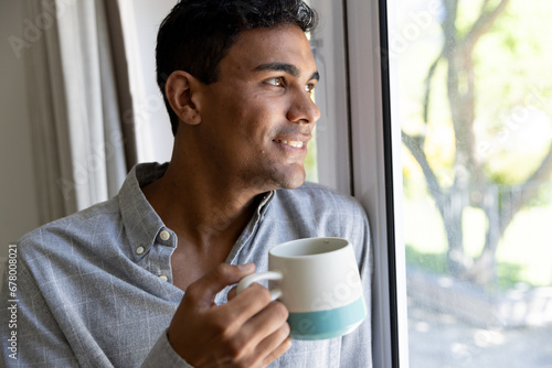 Happy biracial man holding mug looking through window at home