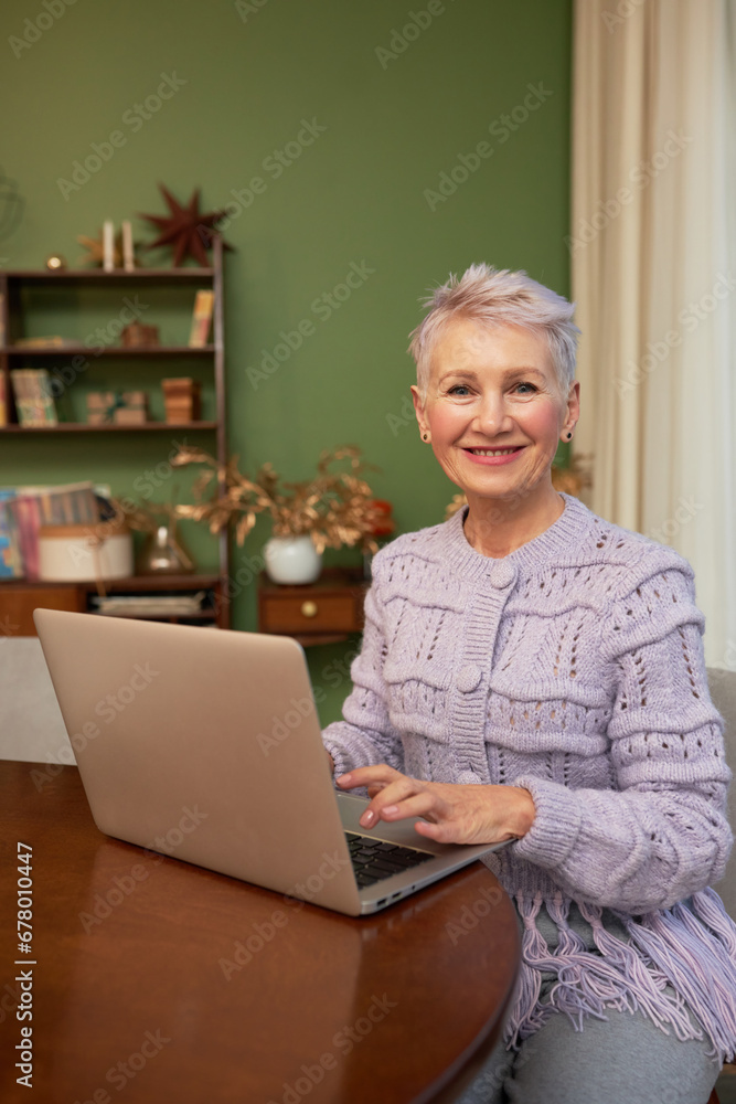 Vertical portrait of smiling senior female using laptop sitting at table in living-room, sending e-mails with christmas greeting cards to her relatives, friends, browsing social media Vertical portrait of smiling senior female using laptop sitting at table in living-room, sending e-mails with christmas greeting cards to her relatives, friends, browsing social media