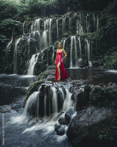 Young Caucasian woman at waterfall in tropical forest. Slim woman wearing long red dress. Beautiful pose. Banyu Wana Amertha waterfall Wanagiri, Bali. Slow shutter speed, motion photography photo
