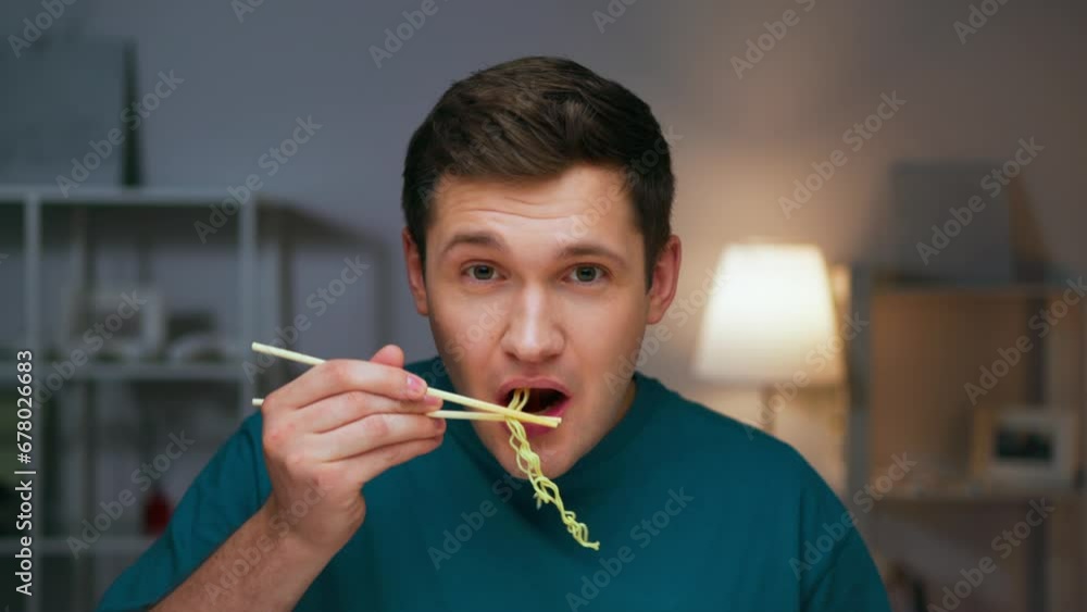 Portrait of Young Man Eating Dinner Food and Looking at Camera Close Up. Modern Person Bites Instant Noodles on Sticks and Relaxes. Appetite of Hunger Guy with Japanese Pasta in Living Room Interior Portrait of Young Man Eating Dinner Food and Looking at Camera Close Up. Modern Person Bites Instant Noodles on Sticks and Relaxes. Appetite of Hunger Guy with Japanese Pasta in Living Room Interior