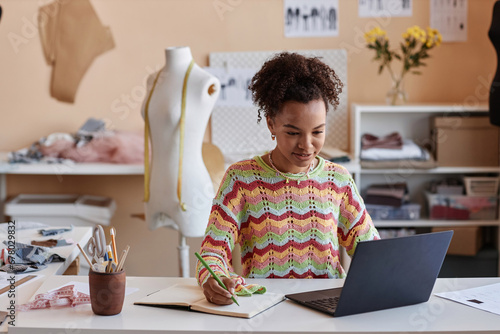 Young African American craftswoman watching online educational video and making notes in copybook while sitting by workplace in craft shop photo