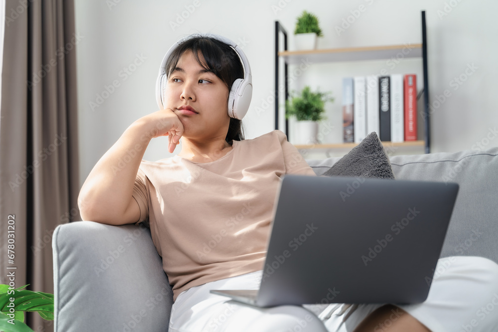 Asian woman wearing over-ear headphones feel bored while studying or working on a sofa in the living room at home. Asian woman wearing over-ear headphones feel bored while studying or working on a sofa in the living room at home.