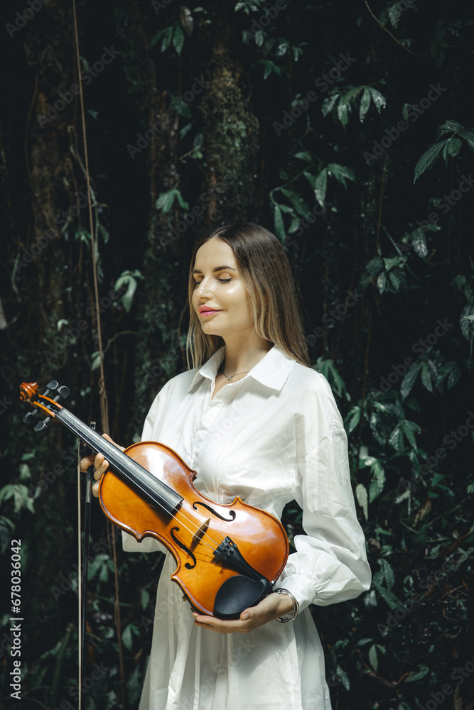 Portrait of charming Caucasian woman holding violin in tropical forest. Closed eyes. Music and art concept. Female wearing white dress. Background of lush green tropical leaves. Copy space.
