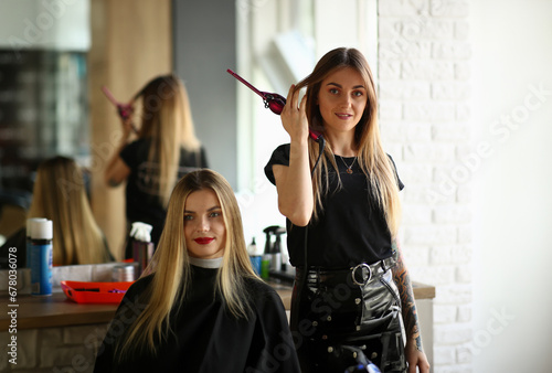Young Girls Hairdresser and Beauty Salon Client. Hairstylist Holding Curling Iron in Hand. Female Customer Sitting in Studio for Making Haircut. Two Beautiful Blonde Woman. Styling Hairdo
