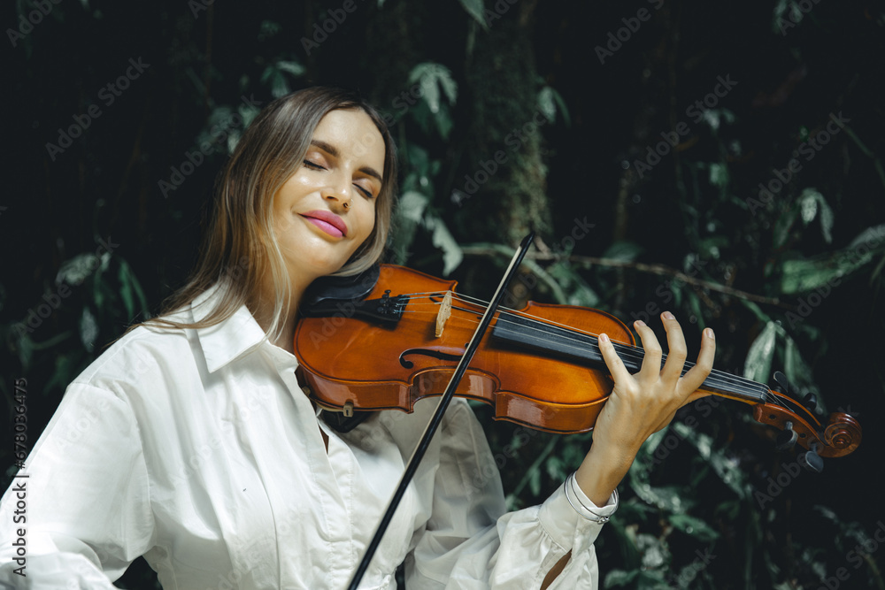 Close up portrait of smiling Caucasian woman playing violin in tropical forest. Closed eyes. Music and art concept. Female with blond hair wearing white dress. Background of tropical leaves.