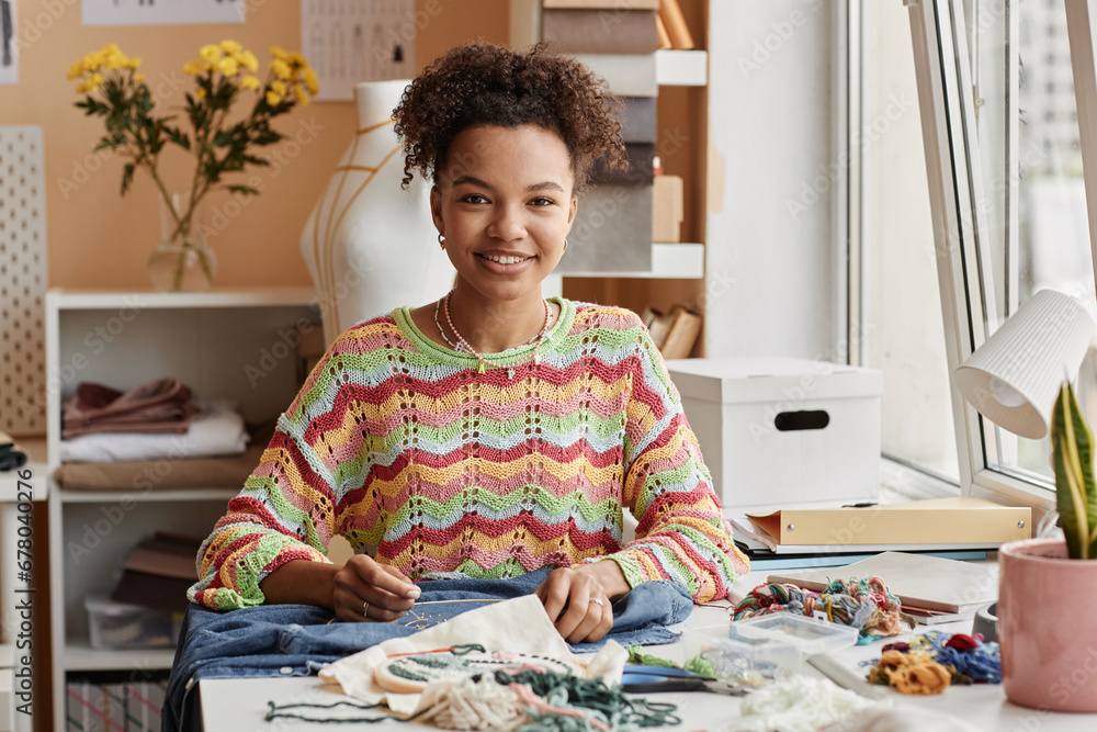 Happy young African American craftswoman looking at camera with smile while sitting by workplace and making beads embroidery on jacket Happy young African American craftswoman looking at camera with smile while sitting by workplace and making beads embroidery on jacket