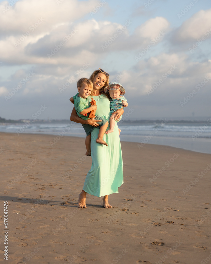 Happy mother walking barefoot on the beach, carrying two sons in her arms. Family relationships. Blue sky with white clouds. Childhood concept. Summer vacation in Asia. Seminyak beach, Bali Happy mother walking barefoot on the beach, carrying two sons in her arms. Family relationships. Blue sky with white clouds. Childhood concept. Summer vacation in Asia. Seminyak beach, Bali