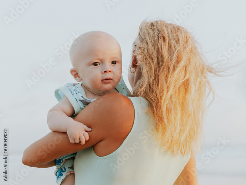 Portrait of Caucasian mother with blond hair and baby son spending time on beach. Summer vacation in Asia. Family relationships. Happy childhood. Sunset time. View from back. Seminyak, Bali