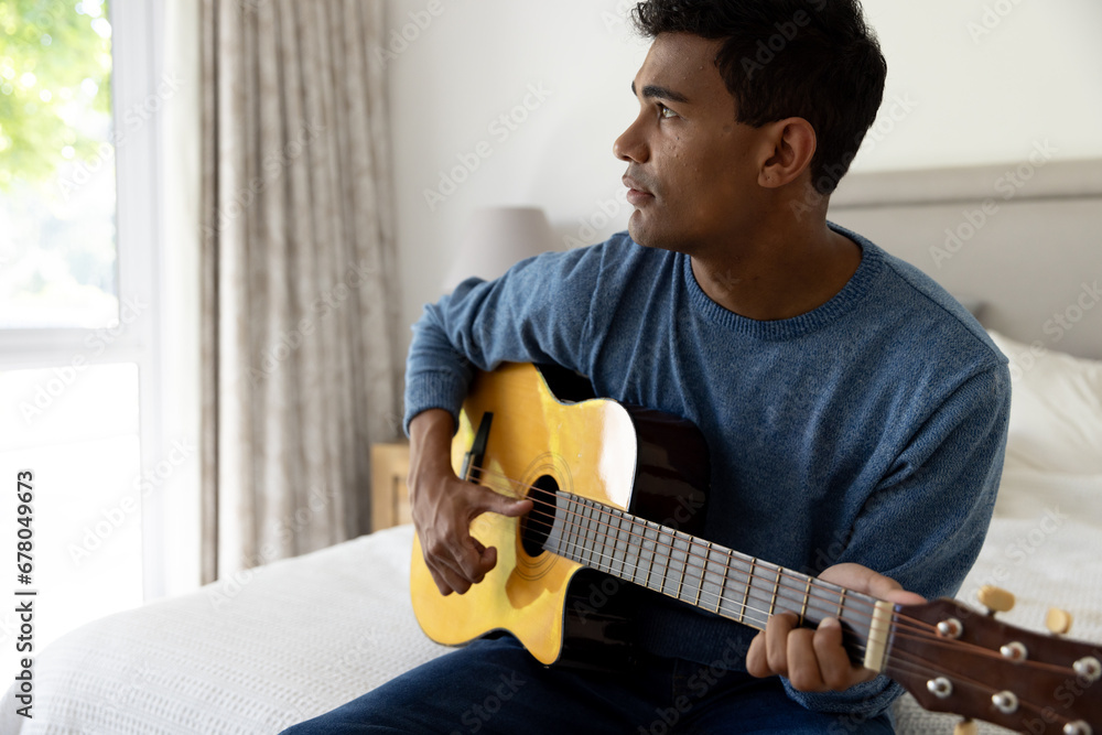 Biracial man sitting on bed playing guitar at home