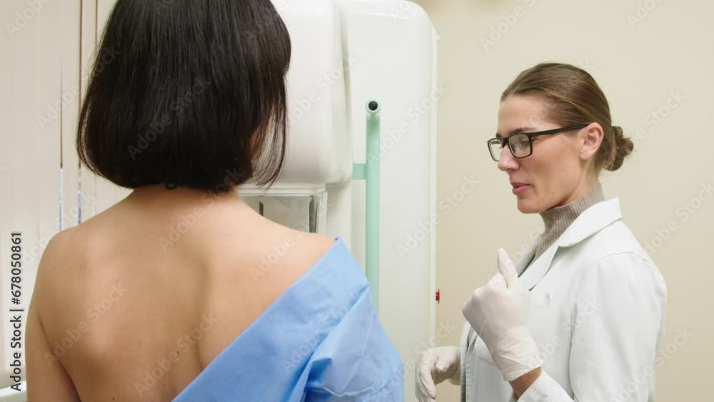 A female doctor in a modern clinic explains the mammography procedure to a topless adult patient undergoing a mammogram. A healthy woman is engaged in cancer prevention in a hospital ward. A female doctor in a modern clinic explains the mammography procedure to a topless adult patient undergoing a mammogram. A healthy woman is engaged in cancer prevention in a hospital ward.