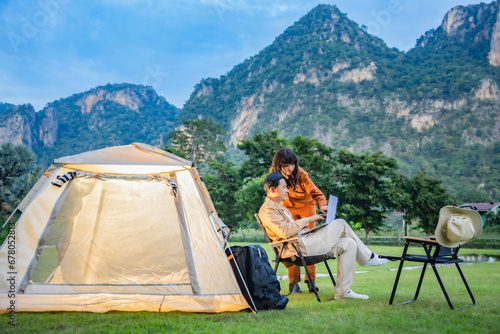 Couple relaxing at camping tent in nature, planning route for leisure travel, searching information on laptop. Mountain valley during sunset. Natural summer landscape on background.