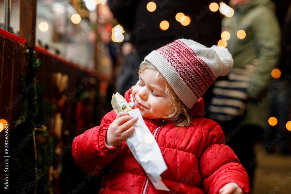 Φωτογραφία Little baby girl, cute child eating bananas covered with chocolate, marshmellows and colorful sprinkles near sweet stand with gingerbread and nuts
