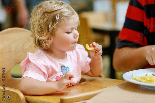 Toddler girl eating healthy vegetables and unhealthy french fries potatoes. Cute happy baby child taking food from parents dish in restaurant. Father eating in fast food restaurant with daughter