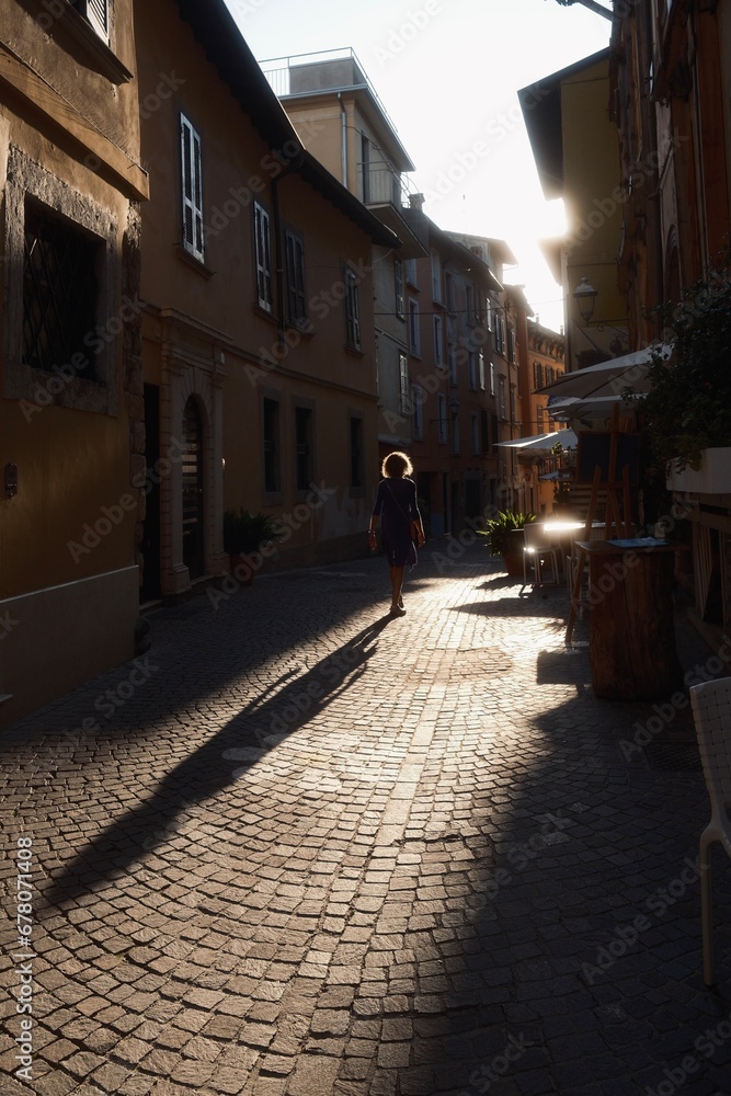 Donna che cammina lungo una via storica con il sole di fronte e ombra molto lunga a terra