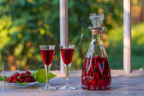 Homemade red raspberry brandy in two wine glasses and in a glass bottle on a wooden windowsill near summer garden, closeup