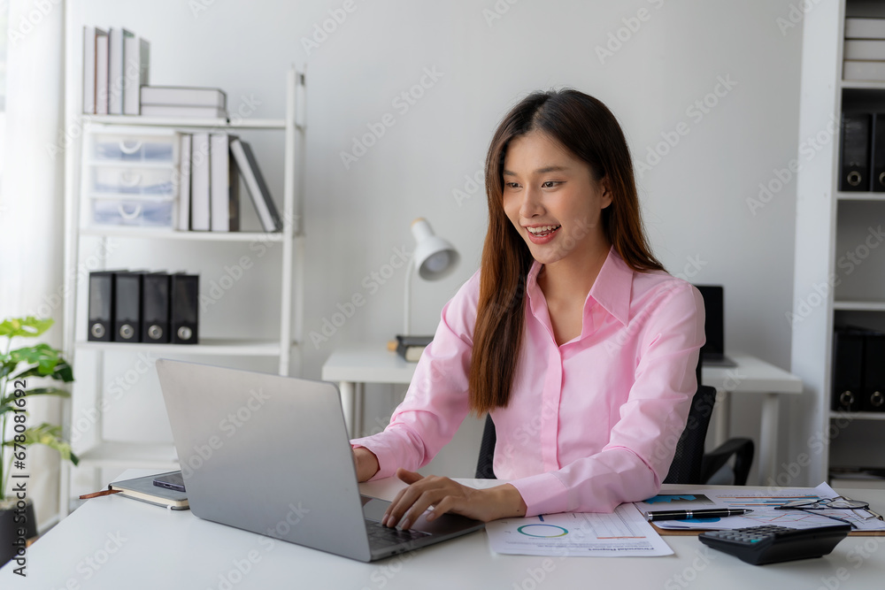 Young Asian woman sitting at desk with data and graph paper using laptop, analyzing accounts, planning strategies on business improvement in office. Young Asian woman sitting at desk with data and graph paper using laptop, analyzing accounts, planning strategies on business improvement in office.