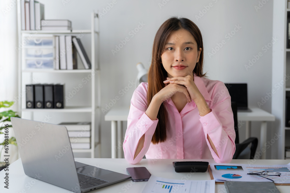 Young Asian woman sitting at desk with data and graph paper using laptop, analyzing accounts, planning strategies on business improvement in office. Young Asian woman sitting at desk with data and graph paper using laptop, analyzing accounts, planning strategies on business improvement in office.